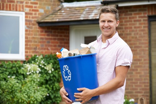 A young man with short light brown hair and a friendly smile is standing outdoors in front of a brick residential building with a white window and a tiled roof. He is wearing a light pink short-sleeved shirt and holding a blue plastic recycling bin filled with various waste items, including paper, cardboard, plastic bottles, and a metal can. The background includes green foliage and flowering bushes, with natural daylight illuminating the scene, emphasizing the context of waste collection and disposal. The image reflects a professional rubbish removal service in Richmond, suitable for both domestic and commercial environments, as delivered by Commercial Waste Richmond. The scene captures a clean, well-maintained environment typical of a suburban property in the postcode area associated with Richmond.