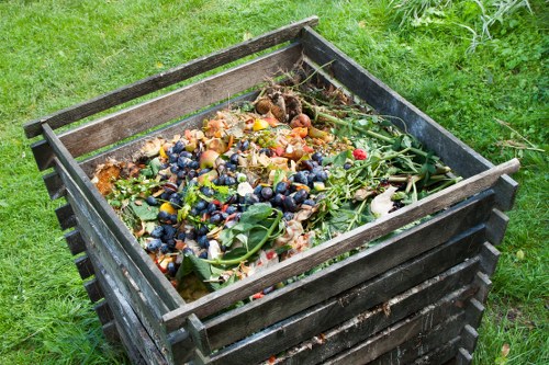 A wooden garden compost bin filled with organic waste including fruit and vegetable peels, leafy greens, and garden clippings, positioned on a grassy outdoor area with green turf surrounding it. The bin is constructed of horizontal wooden slats with a dark finish and metal corner reinforcements, showing signs of use. The organic waste inside appears partially decomposed with a mixture of fresh and slightly decayed plant matter, some colorful berries, and leafy stems visible on the surface. The environment suggests a residential garden setting, aligning with waste collection and rubbish removal services in Richmond, where Commercial Waste Richmond operates. The scene is illuminated with natural daylight, highlighting the textures of the organic materials and the wooden structure of the compost bin.